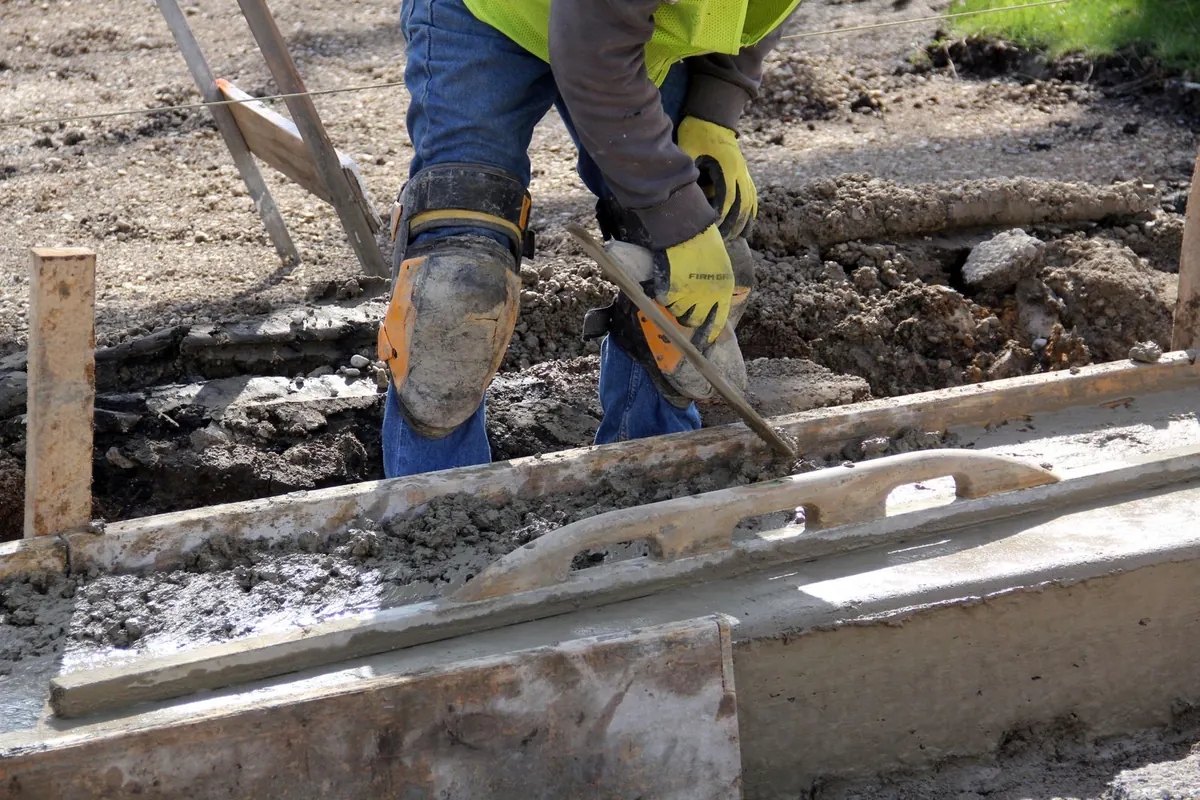Worker leveling wet concrete between wood forms with a screed board