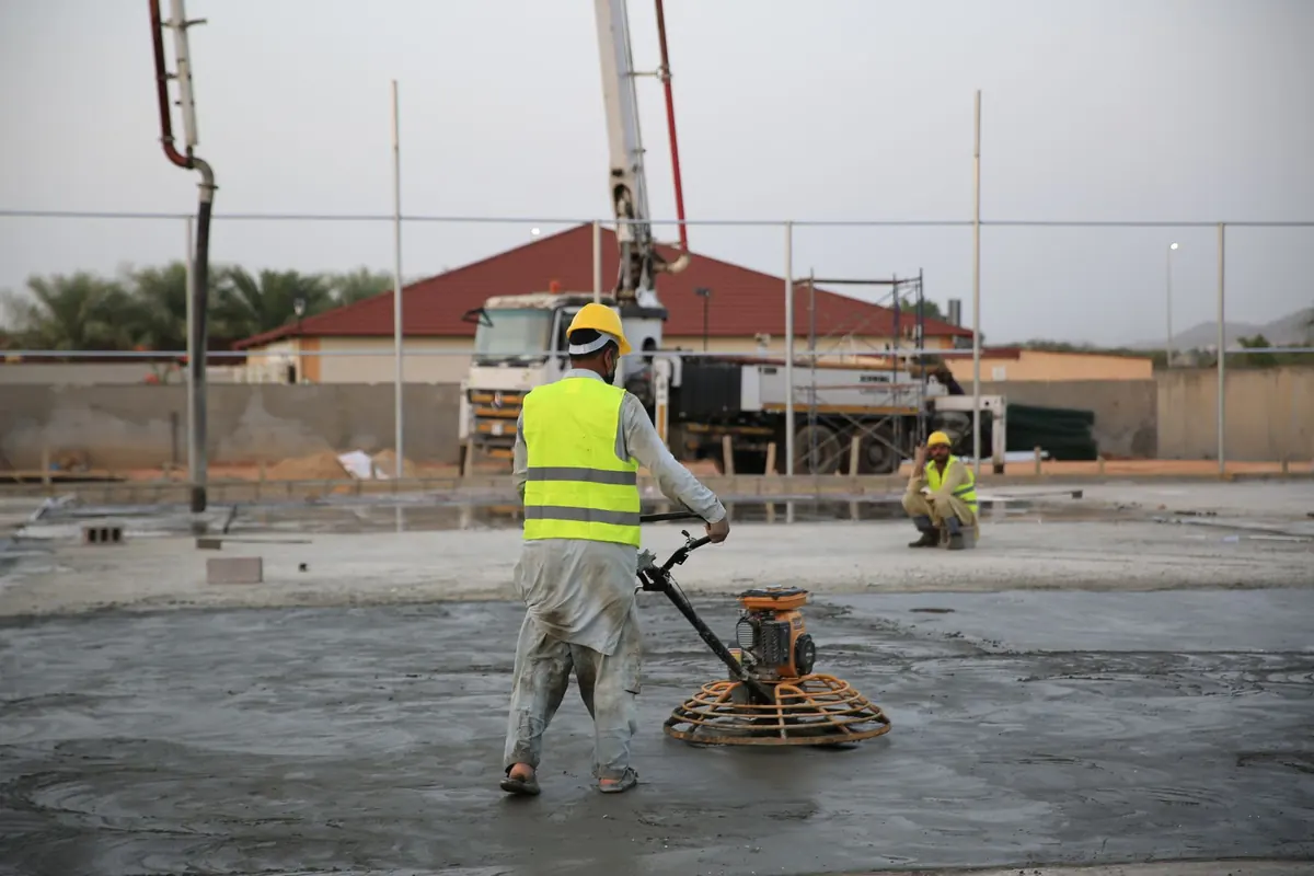 Worker finishing a large concrete slab with a walk-behind power trowel near a pump boom